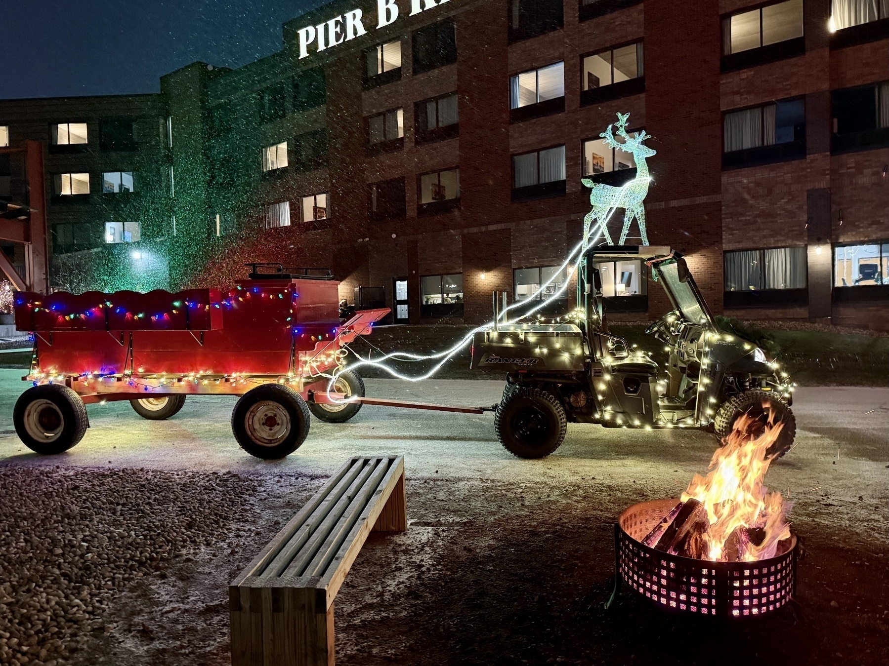 a wagon hitched to an ATV, illuminated in holiday lights, behind a firepit and in front of a building called Pier B Resort, in the evening