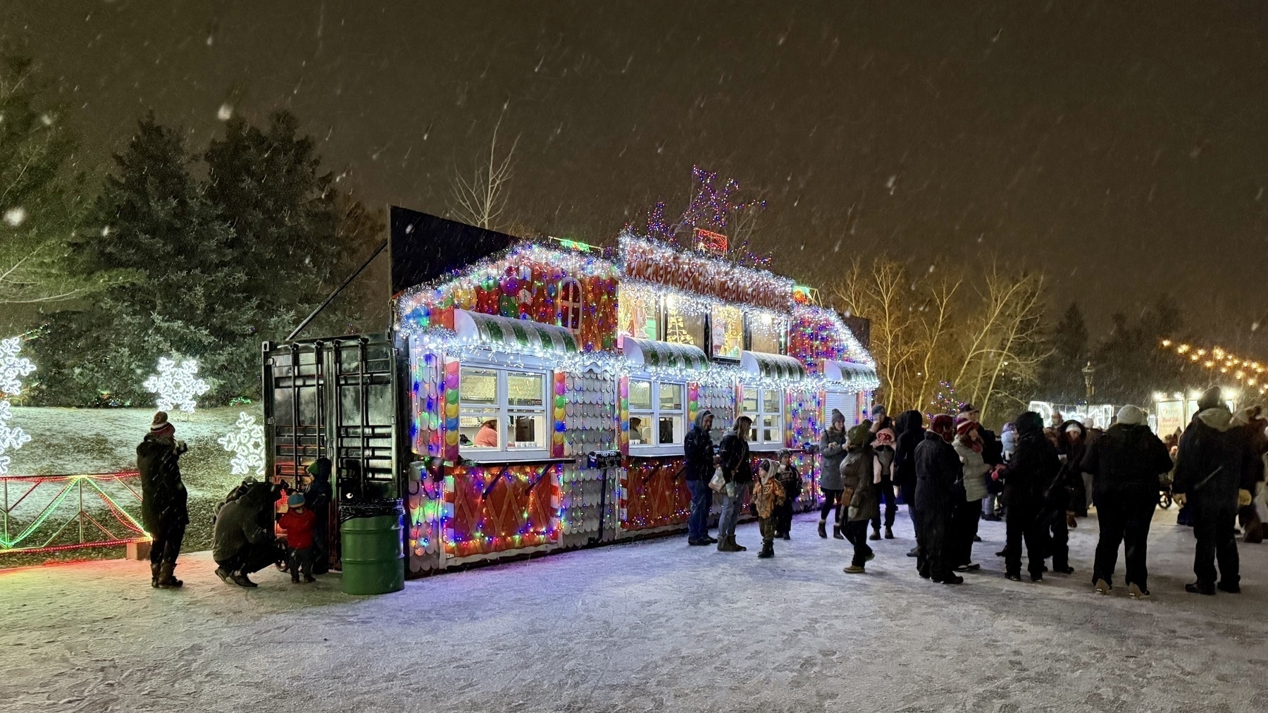 Night view of a holiday-light illuminated outbuilding called Cookie House with a line of folks waiting to get snacks